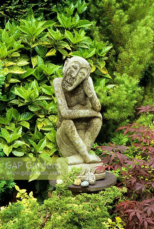 La sculpture en pierre patinée de Bali est davantage mise en valeur en étant entourée de conifères et élevée sur un socle de pot de cheminée coiffé d'un morceau rond d'ardoise - Acer palmatum 'Atropurpureum' en premier plan