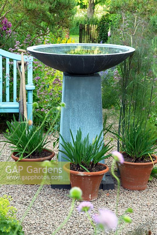 Pièce d'eau contemporaine en pierre bleue avec bol dans le jardin sur gravier avec des pots d'Agapanthus. Banc de jardin bleu avec porte au-delà