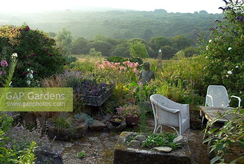 Coin salon avec lavabos alpins, plantes auto-ensemencées, Rosa 'Roserie de L ' Hay' et jardin marécageux avec statue et obélisque. Vue sur la montagne Dinas et le pic Garn Fawr au loin
