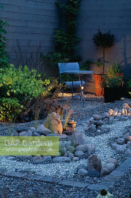 Jardin de nuit avec une rivière de gravier réfléchissante qui court autour de galets, table et chaise en métal en arrière-plan