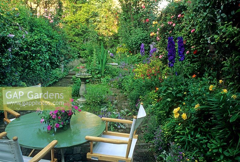 Petit jardin de style cottage de ville avec table et chaises de direction sur terrasse en brique. Delphinium, Campanula, Alchemilla mollis et Roses en parterre de fleurs. Impatiens en pot sur table - Londres
