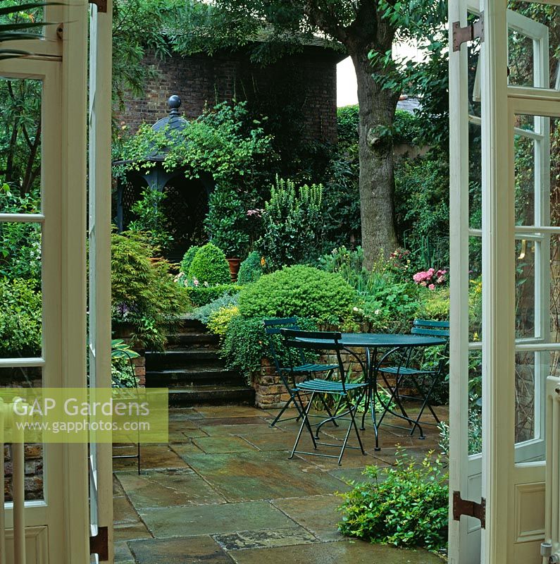 Vue à travers les portes-fenêtres sur la cour pavée, la salle à manger et les marches flanquées d'un feuillage vert luxuriant - Conçu par Jill Billington