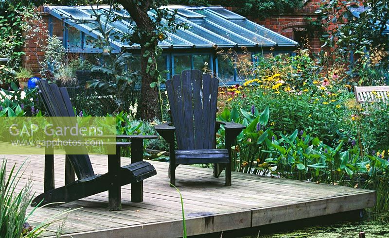 Terrasse en bois à côté de l'étang avec chaises adirondacks et serre en arrière-plan - Greystone Cottage, Oxfordshire