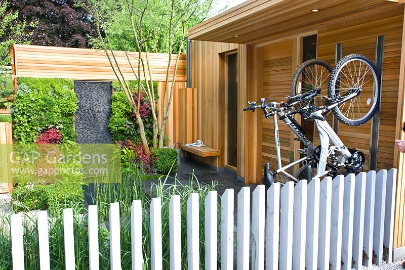 Des vélos dans un porte-vélos dans un jardin moderne avec un mur végétal vivant avec des plantes, y compris des fougères et Heuchera à côté d'un mur d'eau vertical en basalte et un bassin profond dans le jardin de la société pour enfants, conçu par Mark Gregory, parrain - La coopérative, Chelsea Flower Show 2008