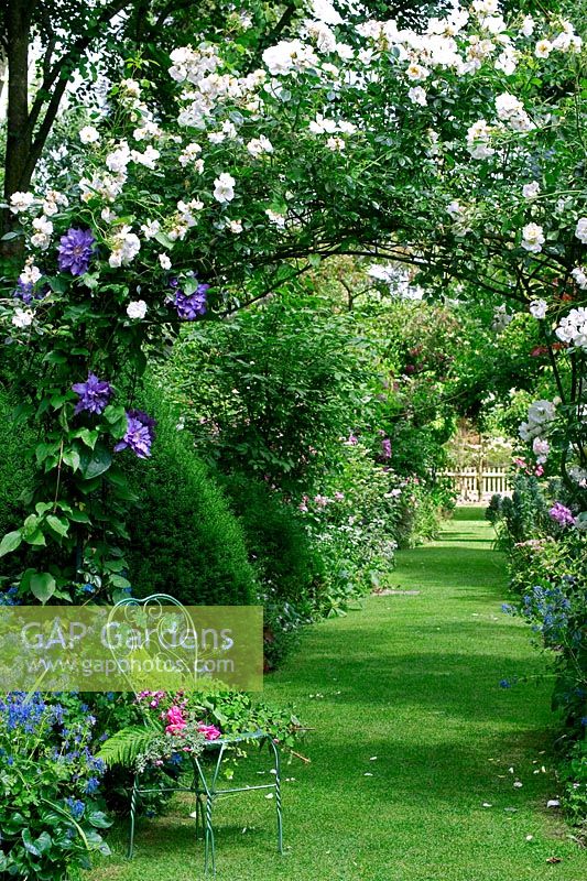 Chemin d'herbe avec siège en métal peint sous une arche couverte de roses - Les Jardins d ' Angélique, France