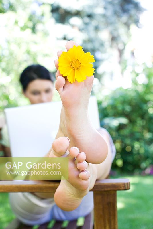 Femme assise à l'aide d'un ordinateur portable sur un banc en bois dans le jardin avec une fleur jaune entre ses orteils