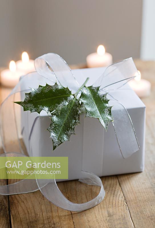 Houx saupoudré de paillettes utilisé pour décorer un cadeau de Noël enveloppé dans du papier blanc et avec du ruban blanc enroulé, avec des bougies en arrière-plan.