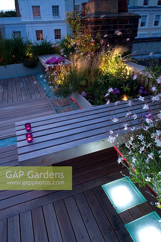 Terrasse en terrasse la nuit avec éclairage led rose et blanc et banc en bois avec bougies, parterres surélevés avec Stipa et Gaura - Roof garden, Holland Park, Londres