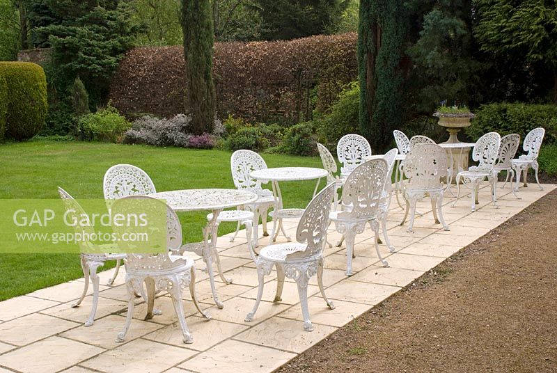 Chaises et tables ornées de métal blanc sur un patio en béton donnant sur la pelouse, la haie de hêtres et le parterre de fleurs avec des conifères et des bruyères - Poulton Hall, jardin NGS, Cheshire