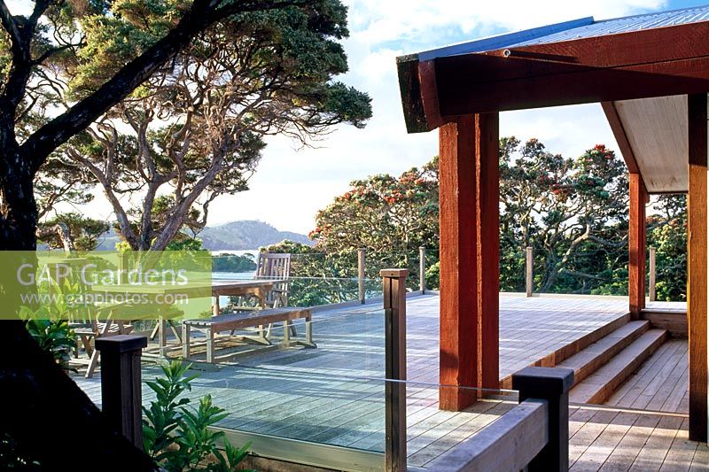Grande terrasse en bois avec garde-corps en verre maximisant la vue mer. Arbres de Pohutukawa et environs de Manuka