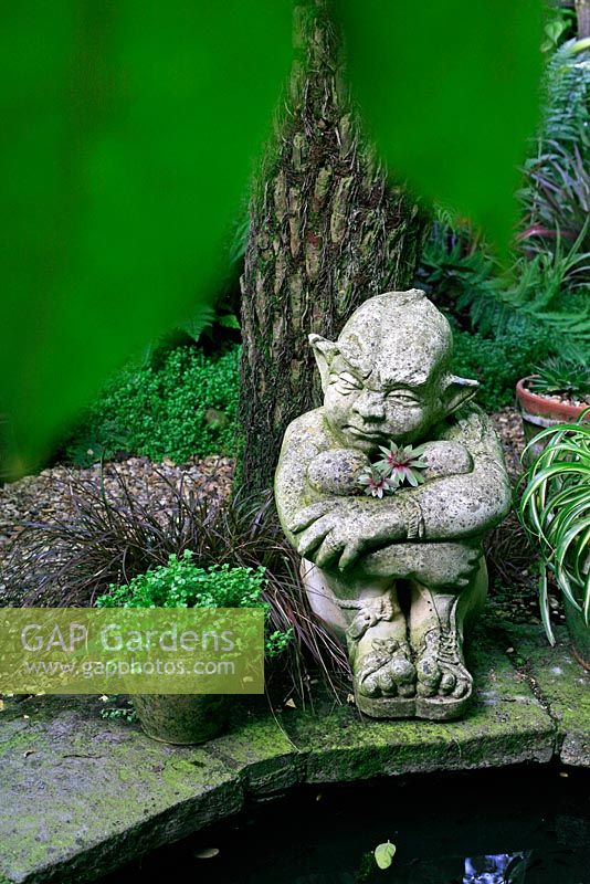 Sculpture d'un lutin assis sous le tronc d'une fougère arborescente à côté d'un étang peu profond avec des joubarbes, Sempervivum jaillissant de son corps
