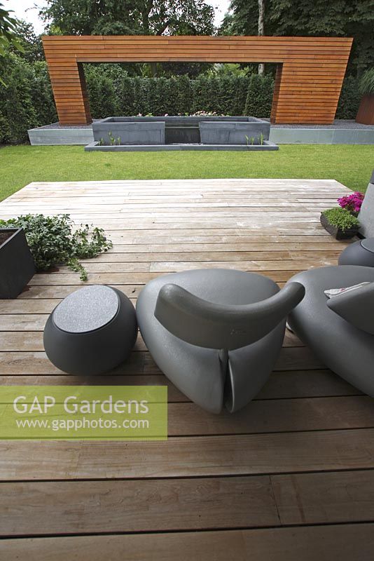 Terrasse dans jardin moderne avec chaises design sur terrasse en bois. Arc en bois artistique avec étang surélevé.