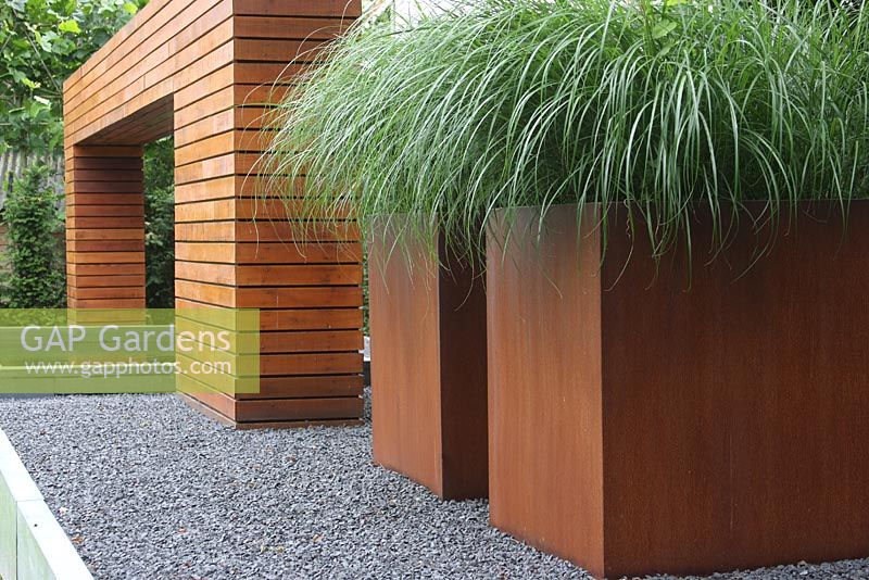 Jardin moderne avec de grandes boîtes rouillées sur un patio surélevé planté d'herbes ornementales. Arche en bois artistique sur l'étang.