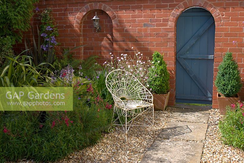 Porte de jardin en bois dans le mur de briques. Dalle de pierre et chemin de gravier avec chaise en fil de fer. 'Hazelwood '. Jacqueline Iddon Hardy Plants, jardin NGS, Lancashire