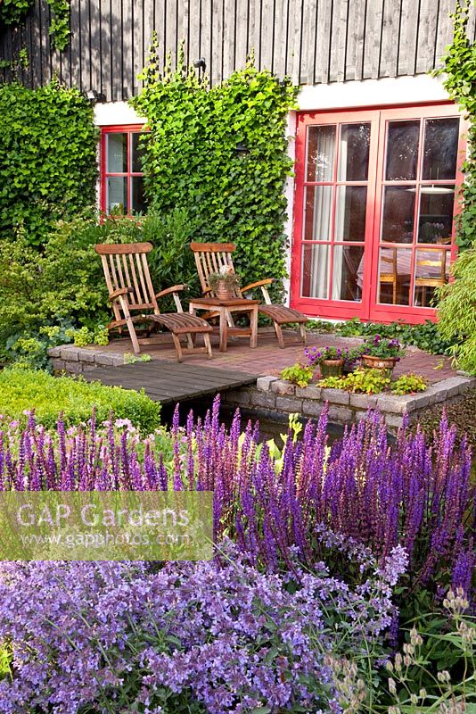 Patio dans jardin paysager avec chaises longues en bois. Parterres de Nepeta nervosa et Salvia nemorosa 'Caradonna'