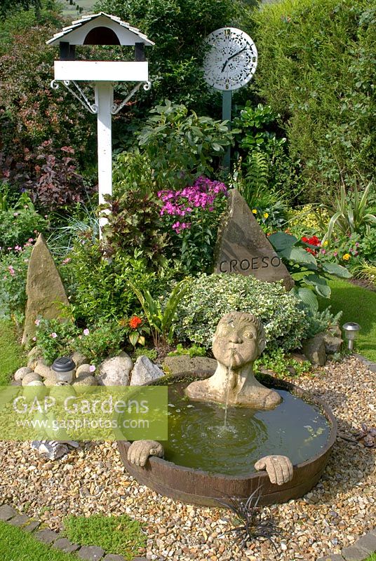 Coin d'un petit jardin à l'arrière avec parterres de fleurs mixtes, haie de Leylandii et caractéristiques comprenant - une table d'oiseau peinte, un élément d'eau à demi-baril avec bec et mains submergés, cadran d'horloge en plateau de table orné peint et sculpture en pierre sculptée avec 'Croeso' - bienvenue en gallois à 'Trevinia', Stubbins, Lancashire NGS