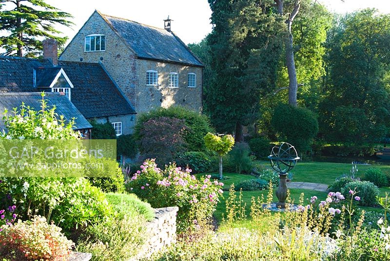 Le jardin supérieur avec cadran solaire et parterres de fleurs en forme d'herbes, de plantes vivaces herbacées, y compris des pivoines et des arbustes intéressants avec étang de moulin vu au-delà - Mill House, Netherbury, Dorset