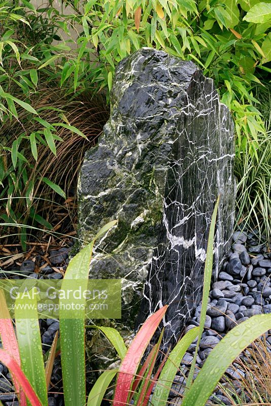 Roche marbrée forée déversant de l'eau à travers un parterre de galets dans un puisard souterrain et entourée de feuillage à feuilles herbeuses, notamment de bambou, de Phormium et de Carex