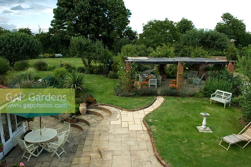 Vue élevée d'un grand jardin de campagne avec pelouse, pergola et coin repas sur terrasse avec marches courbes. Parsons Cottage, Essex, Royaume-Uni
