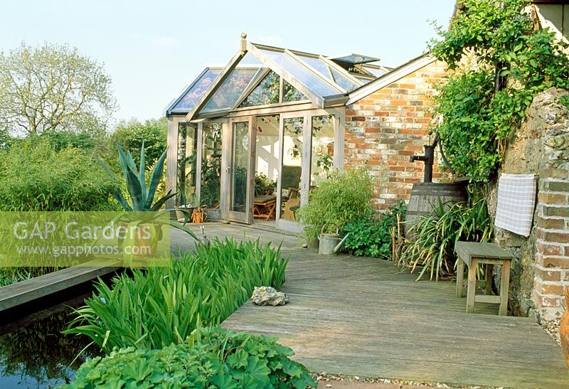 Terrasse en bois, étang et véranda. Banc en bois et Agave en pot. Fin du printemps, Fovant Hut Garden, Wilts