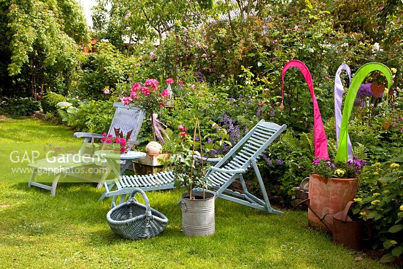 Chaises longues sur la pelouse à côté de parterre de fleurs avec Rosa et ornement coloré fait de bandes de tissu en pot en terre cuite