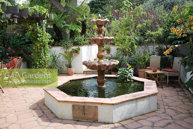 Fontaine à gradins en marbre italien en piscine surélevée octogonale sur carreaux de terre cuite. Vigne sur pergola en chêne et chaises en osier dans un petit jardin urbain