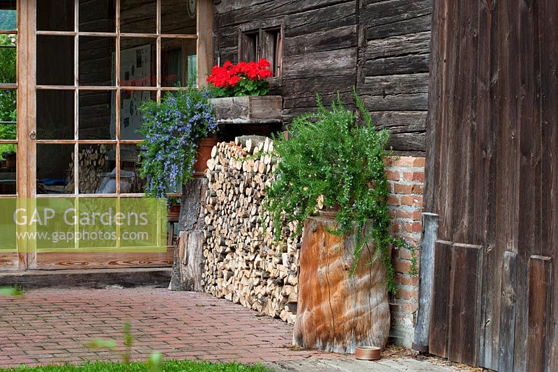 Entrée de la maison avec porte en verre, pile de bois, une fenêtre avec Zonale Pelargonium et Rosmarinus officinalis