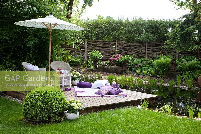 Mobilier de jardin en osier blanc, une couverture avec des coussins et un parasol sur une terrasse en bois à côté d'un étang de jardin. Treillis de saule et haie en arrière-plan. Les plantes sont coupées Buxus, fougère et hortensia
