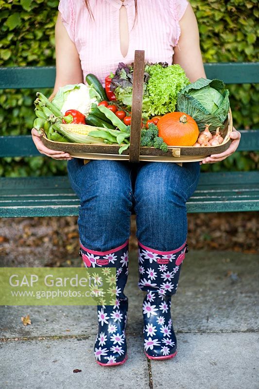 Femme portant des jeans et des bottes en caoutchouc assis sur un banc de jardin tenant un trug en bois de légumes d'automne