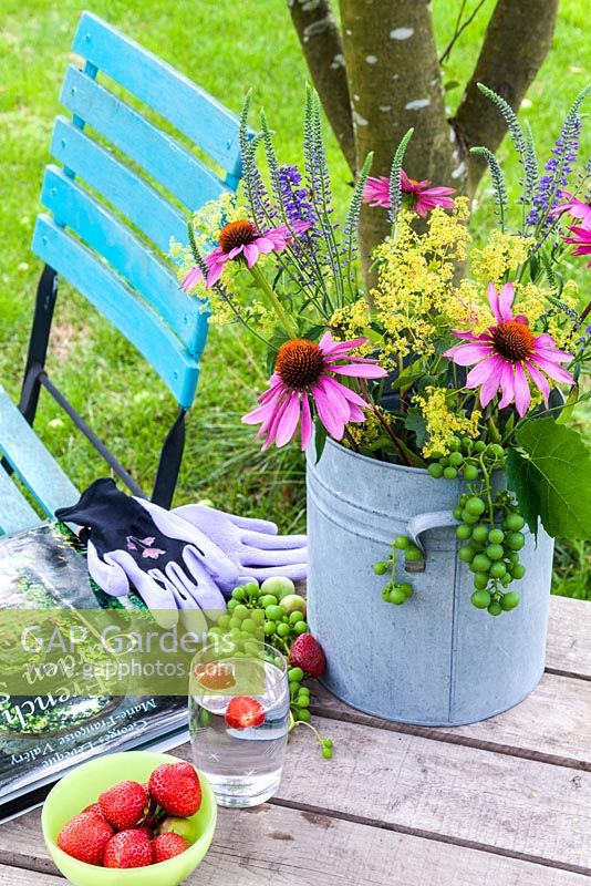 Sur une table en bois un vase avec coneflower, Ladys Mantle, Veronica et raisins. Également sur la table, un livre de jardinage, des gants de jardinage, une fourchette à main, un bol de fraises et un verre d'eau. En arrière-plan une chaise de jardin turquoise colorée.