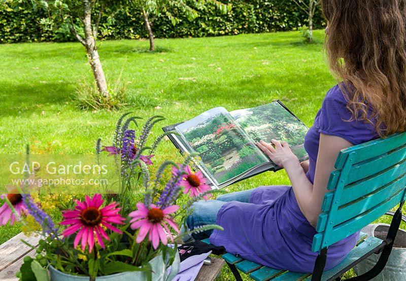 Femme portant une robe d'été violette assise sur une chaise turquoise lisant un livre de jardinage à l'ombre. Au premier plan un vase avec coneflower, Ladys Mantle et Veronica. Sur la table en bois des gants de jardinage.
