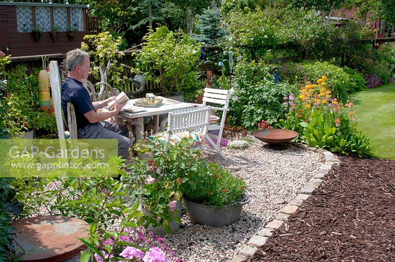 Homme assis dans un coin gravelé de jardin avec petite table et chaises et collection de pots galvanisés d'époque - la plantation comprend; Rosa, Primula, Argyranthemum et Geranium
