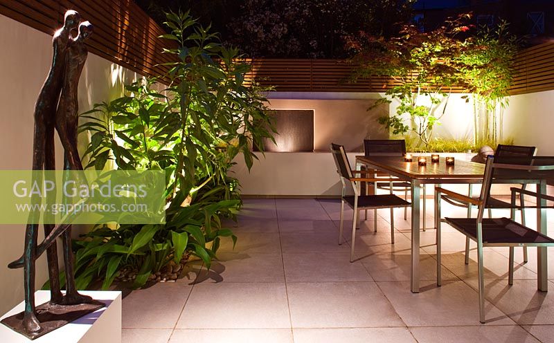 Jardin minimaliste éclairé la nuit avec table et chaises - la plantation comprend Beschorneria Yuccoides 'Quicksilver'