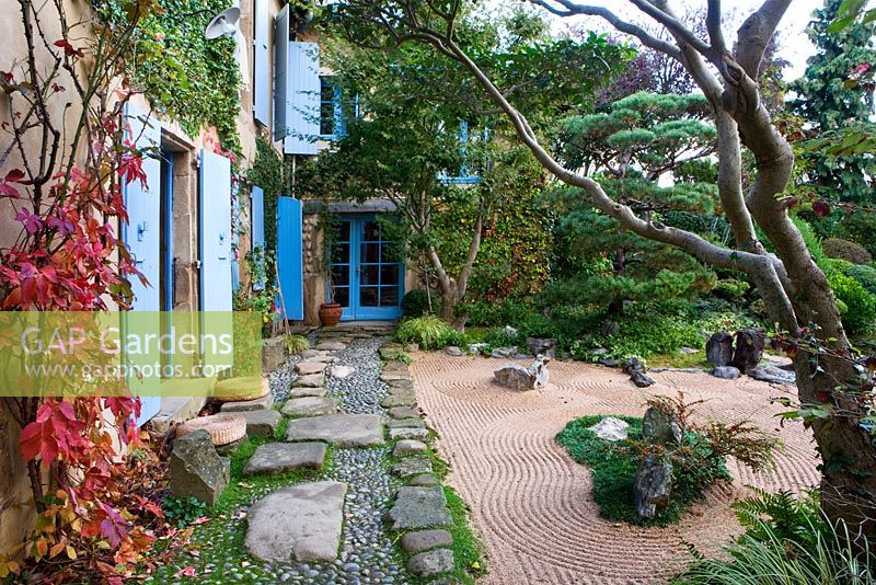 Jardin de style japonais - vue sur le côté est de la maison avec volets bleus, chemin avec galets et rochers, jardin de gravier et pin sylvestre taillé