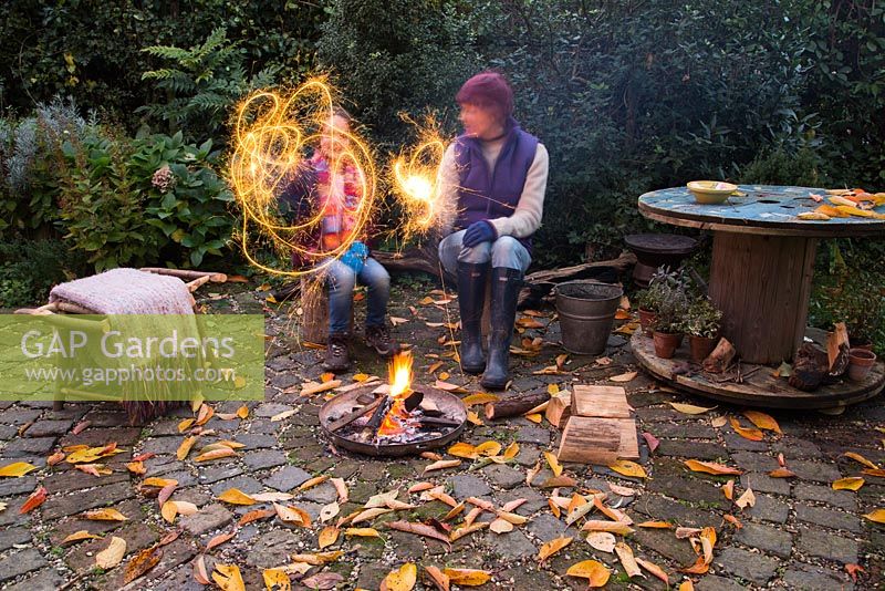 Mère et fille allumant des cierges magiques par un foyer dans un jardin arrière automnal.