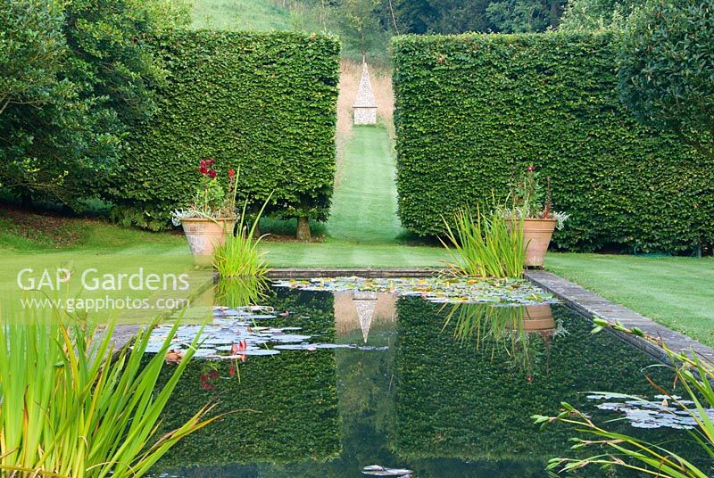 Étang formel bordé de Crataegus x lavallei standard dans le jardin supérieur où un espace entre des haies de hêtres encadre un obélisque situé dans une prairie herbeuse. Corscombe House, Corscombe, Dorset, Royaume-Uni