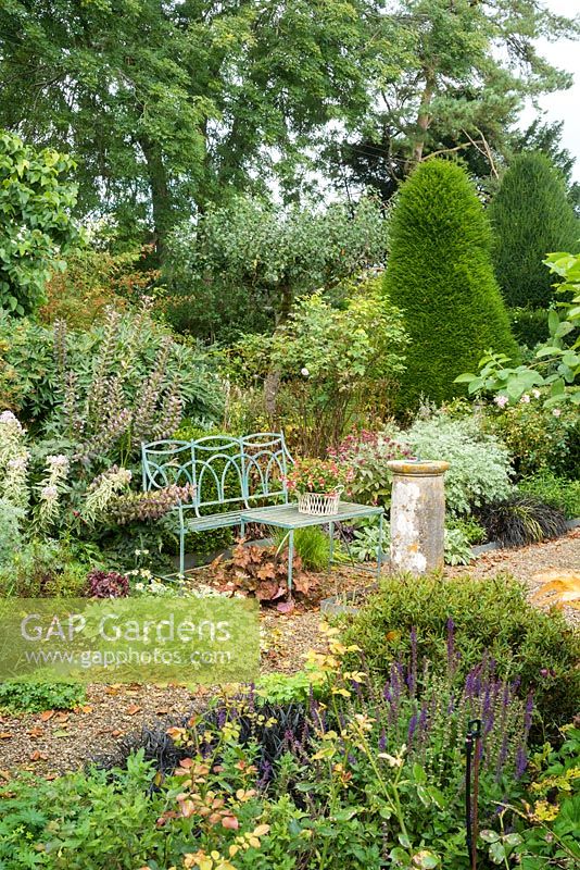 Jardin à la fin de l'été avec banc en fer forgé peint en bleu, vieux cadran solaire, chemins de gravier, roses, plantes vivaces herbeuses et vue sur l'ifiaire.