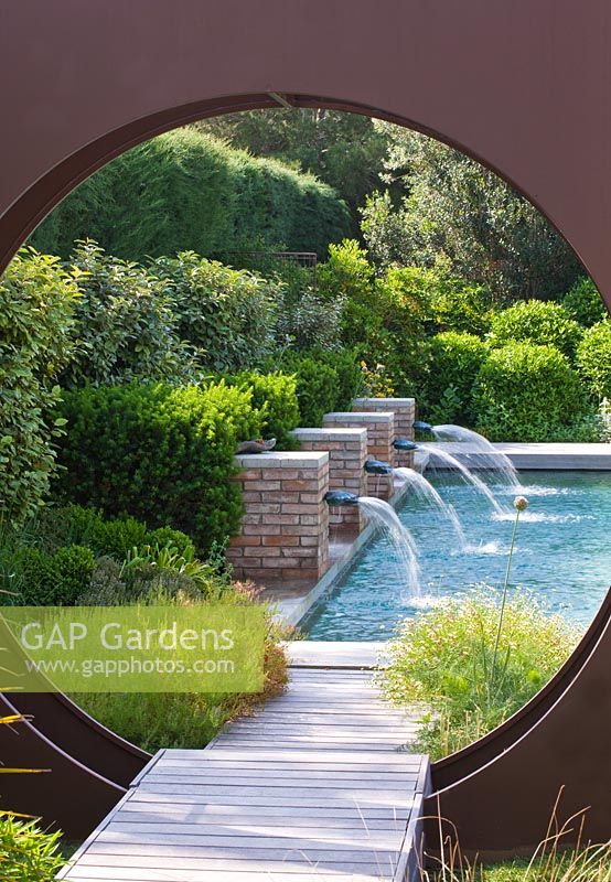 Jardin contemporain moderne - cercle 'oculus' en métal et piscine au-delà avec quatre jets d'eau en brique