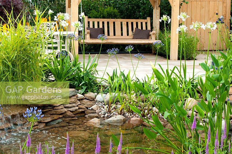 Piscine bordée de galets et de galets adjacente à la terrasse et au siège avec plantation d'Agapanthus et de Sagittaria dans The Water Garden - RHS Tatton Park Flower Show 2013.