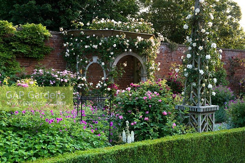 Obélisque en bois et siège couvert de tonnelle en bois dans un jardin clos. Parterres de fleurs aux bords plantés de roses et de psilostemon géranium. Seend, Wiltshire