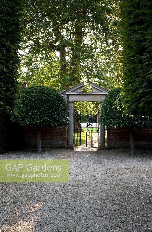 Vue depuis l'allée de style français à la porte formelle dans le jardin clos avec fronton en pierre ornée et arbres topiaires symétriques jumeaux - Seend, Wiltshire
