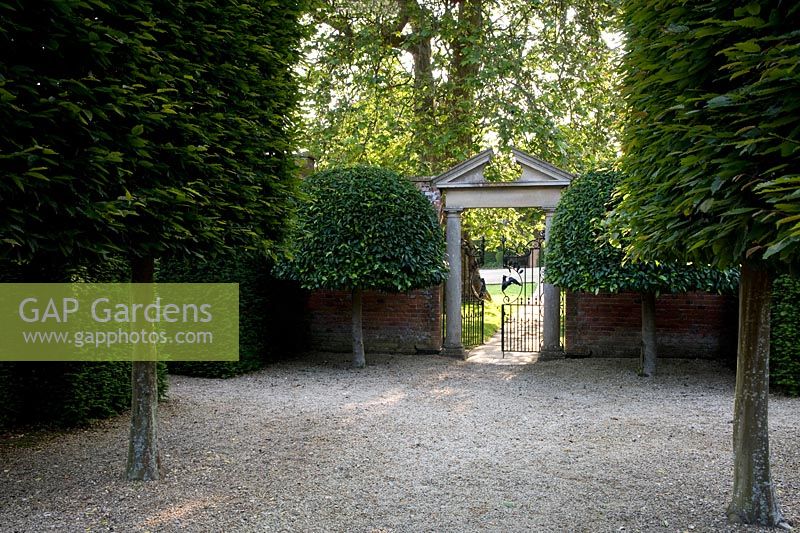 Vue depuis la grande allée de style français à la porte formelle dans le jardin clos avec fronton en pierre et arbres topiaires jumeaux de chaque côté. Seend, Wiltshire