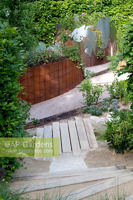Titre: Paradigme. Petit pont en bois comme entrée du jardin Paradigme avec des baignoires ovales en fer rouille remplies de plantes.