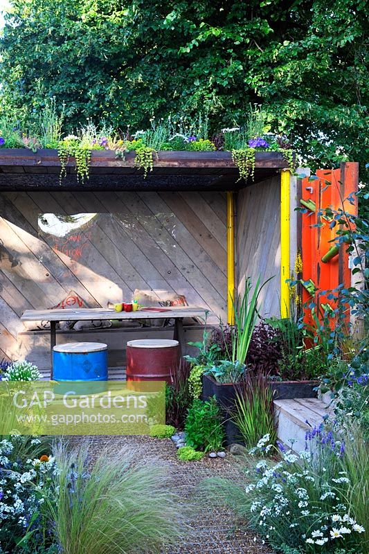 Un espace pour se connecter et grandir. Espace abri en bois avec toit végétal vivant, tabourets de baril de pétrole recyclé, table en bois, élément d'eau en bambou, plantation dans un abreuvoir et siège en bois recyclé.