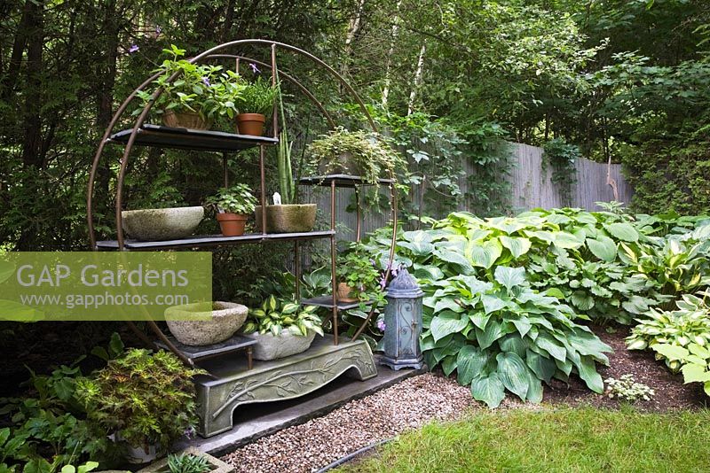 Tube circulaire en cuivre et étagère murale en métal décoré de plantes annuelles et vivaces en pot dans un jardin paysager en été. Rangée de plantes lys 'Hosta' à droite - Il Etait Une Fois, Québec, Canada.
