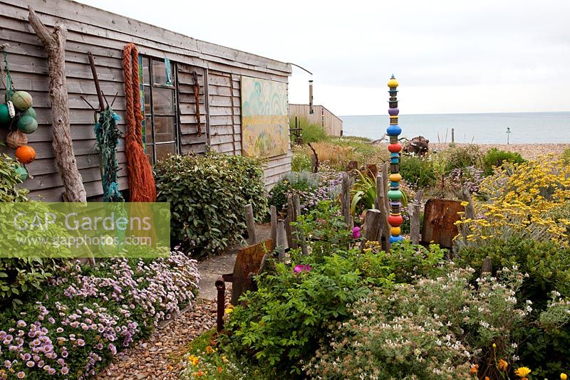 Jardin côtier avec cabanon rustique, filets, bouées, sculpture peinte, bois flotté, erigeron et santolina
