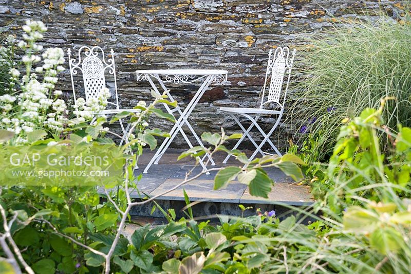 Table et chaises de café en métal contre un mur de pierre, encadrées de miscanthus et d'autres arbustes.