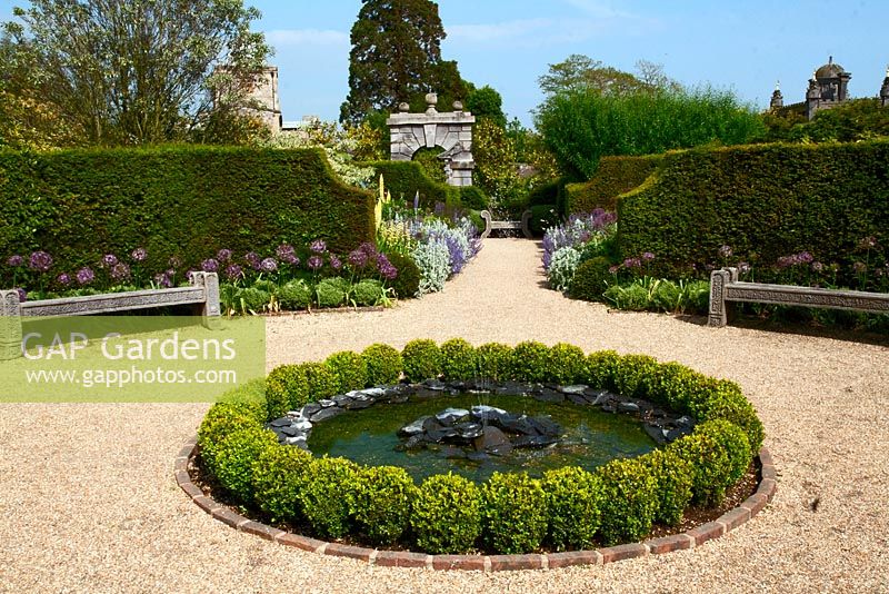 Château d'Arundel - étang circulaire avec fontaine bordée de boules Buxus, bancs en pierre, Allium et parterres de fleurs herbacées jumelles au loin