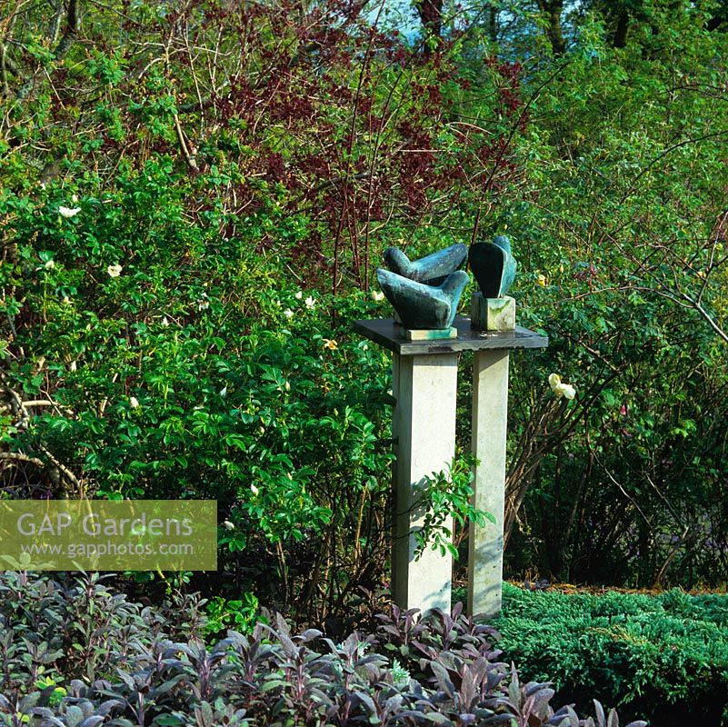 Un socle s'élève sur un fond de sauge, de genévrier et de roses, portant 'Trois colombes cycladiques' de Bridget McCrum.