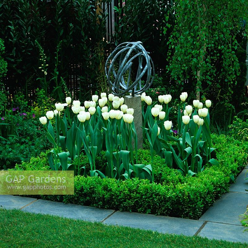 Parterre de fleurs carré formel, entourage pavé, avec boîte entourant des tulipes blanches s'élevant autour de la sculpture moderne.